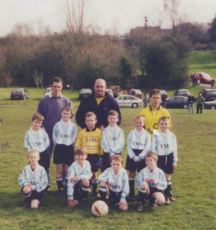 The first ever Fleur-De-Lys U7 Football Team.

A group photograph of a youth football team, consisting of players wearing light blue and white jerseys with 'TMS' printed on them, posing together on a grassy field with two coaches in the back. The setting includes some parked cars and trees in the background.